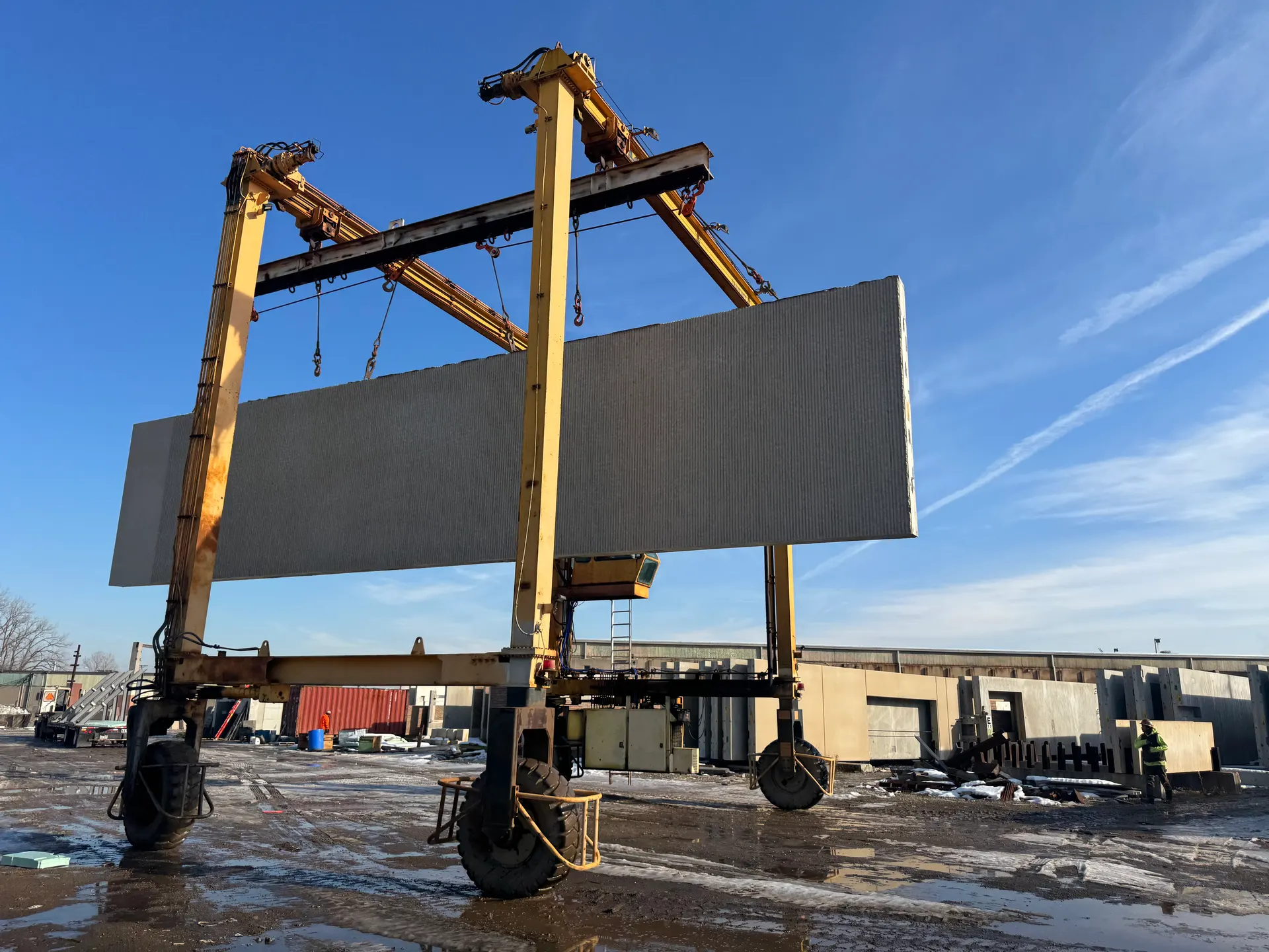 A straddle carrier lifts a long precast wall panel across a muddy plant yard under a clear winter sky.